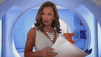 A woman with light brown hair is standing in a futuristic blue room, wearing a gray sleeveless top and holding papers. She has a serious expression.